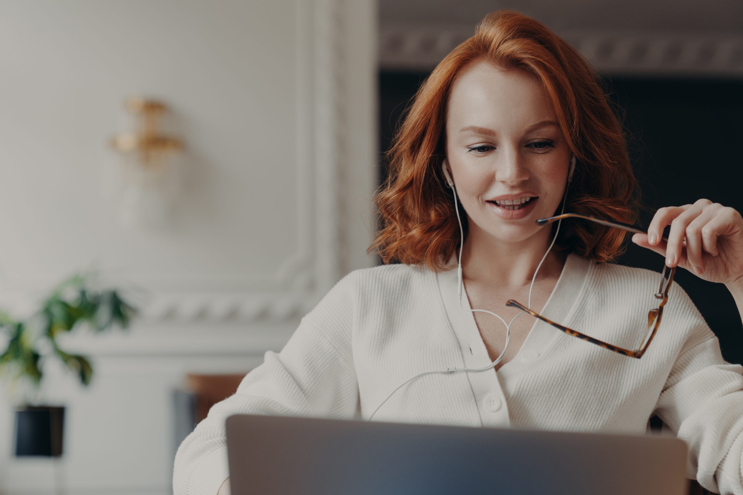 Pleased redhead woman student watches training webinar, has online video conference, uses earphones, works on business project, keeps transparent glasses in hand. Distance work via internet.