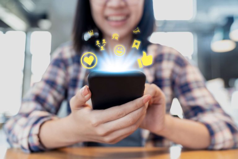 A smiling woman holding a smartphone with glowing social media icons—such as a heart, thumbs up, and speech bubble—floating above the screen.